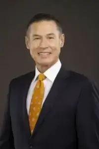 square_4_media A man wearing a dark suit, white dress shirt, and patterned orange tie poses in front of a plain dark background, smiling at the camera. media_agency