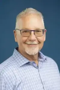 square_4_media A middle-aged man with short gray hair, glasses, and a trimmed beard smiles at the camera, wearing a blue and white checked shirt, against a solid blue background. media_agency