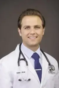 square_4_media A man in a white lab coat with a stethoscope around his neck poses for a professional portrait against a plain background. media_agency