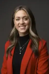 square_4_media A woman with long, highlighted hair wears a red blazer over a black top, layered necklaces, and smiles at the camera against a dark background. media_agency