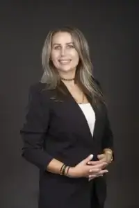 square_4_media A woman with long, light brown hair wearing a black blazer and white top stands against a dark background, smiling at the camera with her hands clasped. media_agency