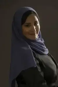 square_4_media A woman wearing a dark purple hijab and black top is looking at the camera and smiling with a neutral dark background. media_agency
