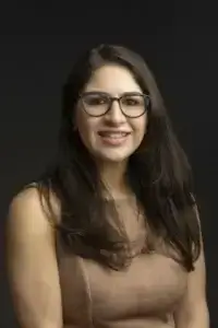 square_4_media A woman with long dark hair and glasses, wearing a sleeveless beige top, smiles while facing the camera against a plain dark background. media_agency