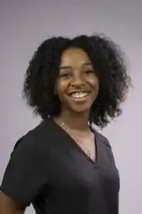 square_4_media A young woman with curly hair smiles at the camera, wearing a black shirt and a necklace, against a plain gray background. media_agency