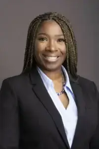 square_4_media A woman with braided hair wearing a black blazer, light blue blouse, and a blue beaded necklace, smiling in front of a plain gray background. media_agency