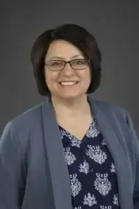 square_4_media Woman with short dark hair and glasses smiling, wearing a patterned navy blouse and a light blue blazer, posed in front of a plain dark gray background. media_agency