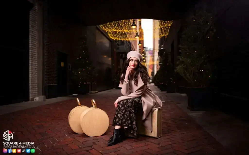 square_4_media A woman in a beige coat and hat sits on a suitcase in a brick alleyway decorated with lights and trees, looking thoughtfully to the side. media_agency square_4_media A woman in a beige coat and hat sits on a suitcase in a brick alleyway decorated with lights and trees, looking thoughtfully to the side. media_agency